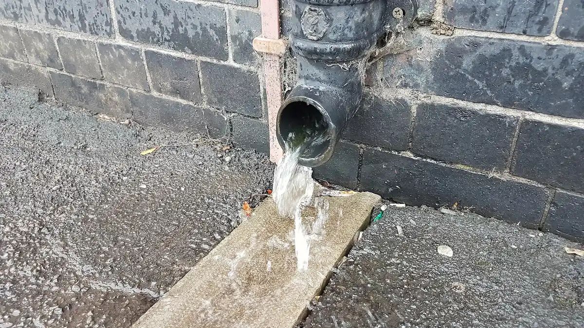 Cast iron downpipe shoe pouring rainwater onto a path beside a brick wall